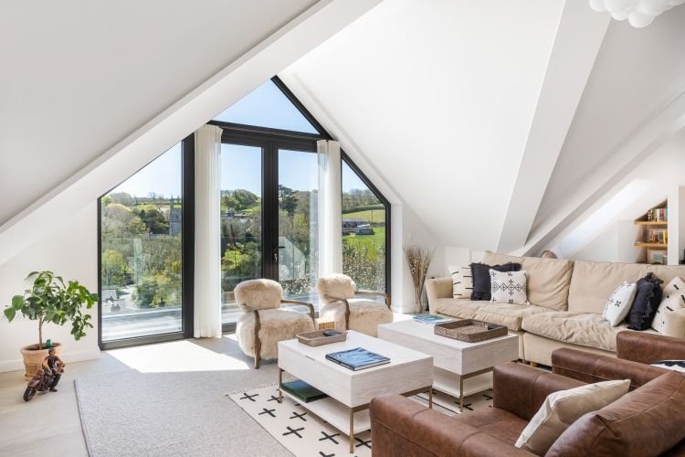 Bright attic living room, captured by a skilled commercial photographer in Cornwall, features a sectional sofa, armchairs, and a coffee table. A large triangular window overlooks greenery, complementing the neutral decor that exudes a cozy, modern feel.