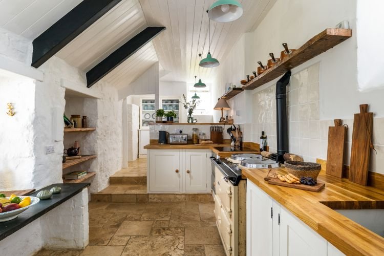 Rustic kitchen with wooden countertops, hanging green lamps, an oven, chopping boards, and stone tile floor. Shelves hold kitchen items, and a fruit bowl is on a side counter.