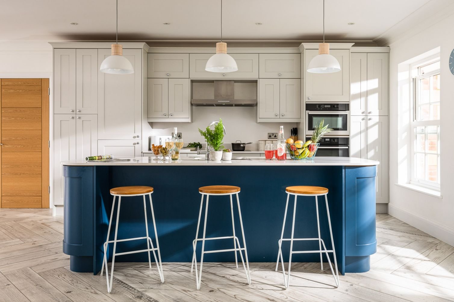 Modern kitchen with a blue island, captured by a commercial photographer in Cornwall. It features three wooden stools, white cabinets, hanging lights, and a warm wooden floor. The island is elegantly decorated with fruits and plants, embodying both style and functionality.