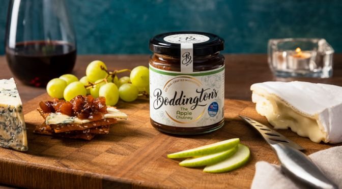 Studio lifestyle product photograph of a cheese board with a jar of chutney. There grapes, apples and a glass of wine in the background.