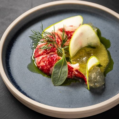 A gourmet dish presents lobster claw, apple slices, green sauce, herbs on a dark ceramic plate.