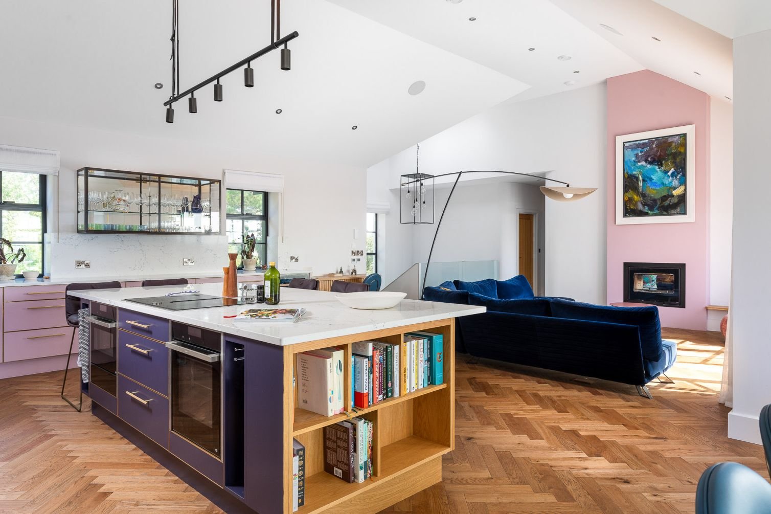 Modern open-plan kitchen and living room boasts herringbone wood floors, navy couches, pink accents, and a large abstract painting above the fireplace.