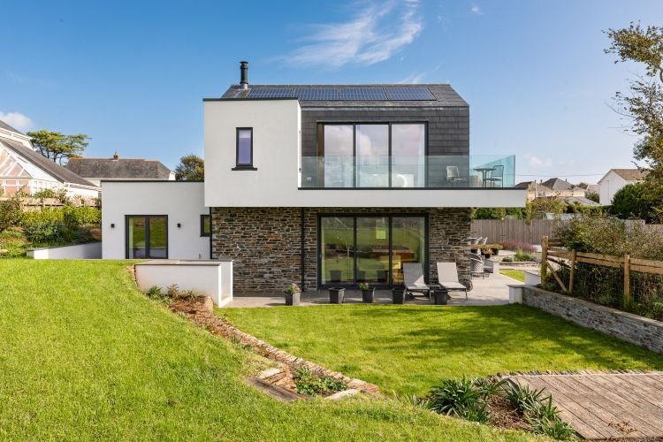 Modern two-story house with stone and white exterior, large windows, a balcony, solar panels, and a grassy backyard with patio and outdoor furniture. An idyllic setting to capture stunning architectural shots.