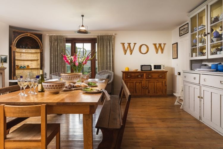 A wooden table is set for a meal, framed by cabinets and a window with curtains. Decorative letters spell "WOW" on the wall, adding charm to this Cornwall setting.