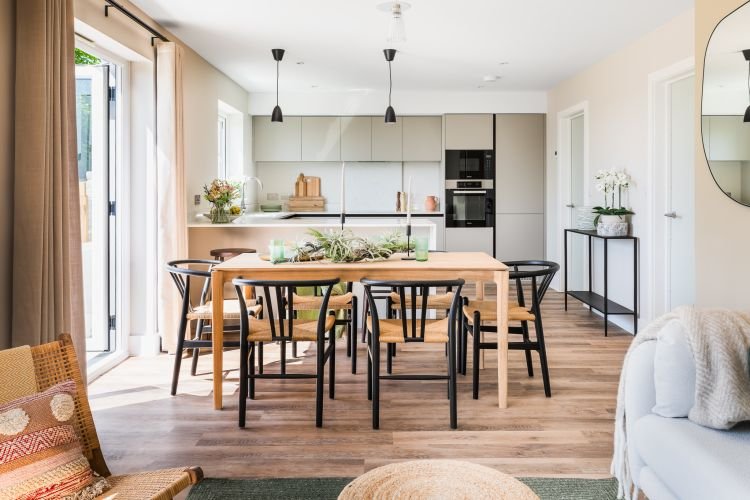 A modern kitchen and dining area with a wooden table, black chairs, and a white and grey kitchen. Natural light floods through the sliding glass doors.