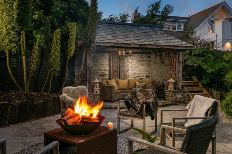 Cozy outdoor seating area with wicker chairs around a lit fire pit. A stone wall backdrop features a sheltered sofa with cushions. Candle and plants add ambiance to the twilight setting.