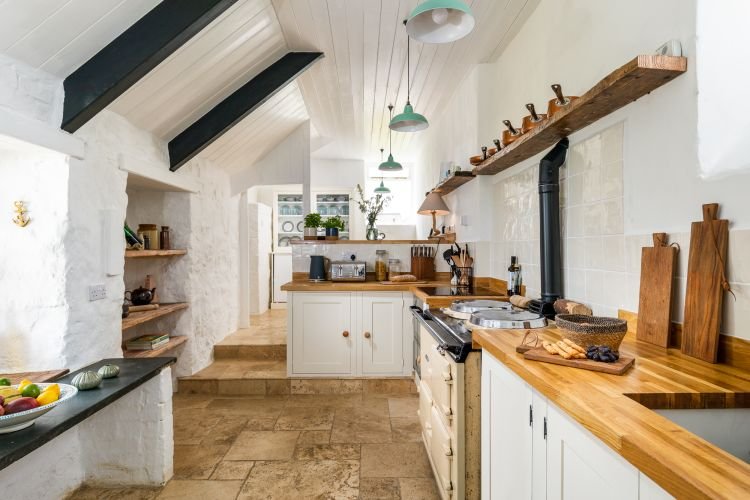 A bright kitchen with wooden countertops, various kitchen utensils, and an oven captures the cozy ambiance. White walls and ceiling, a natural stone floor, and shelves displaying items make it perfect for an interiors photographer in Cornwall seeking charm.