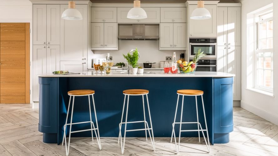 Modern kitchen with a blue island, white cabinets, and three wooden stools captured by an interiors photographer. Pendant lights illuminate the space, with a window providing natural light. The countertop features plants, fruit, and drinks for a touch of freshness.