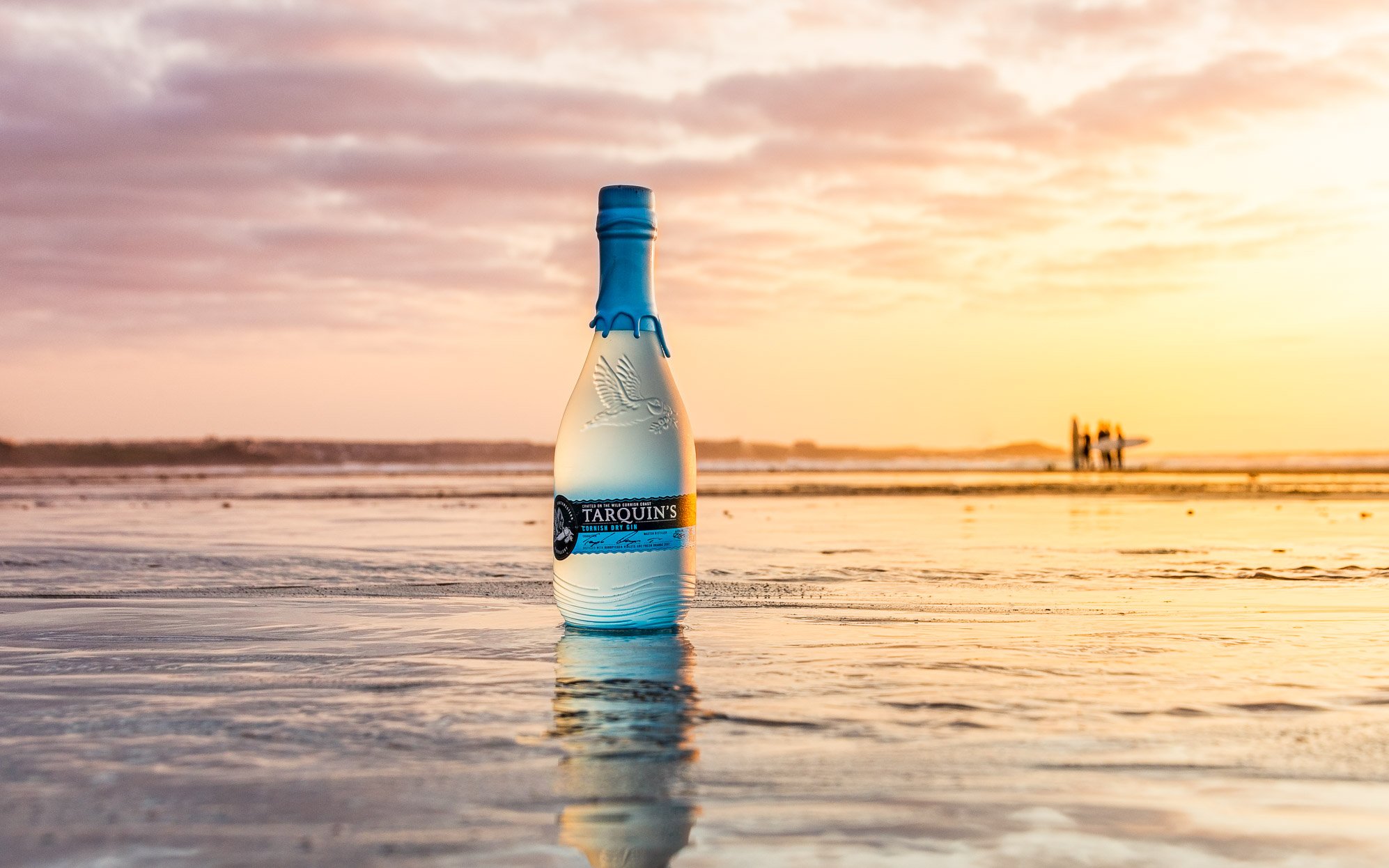 A bottle of Tarquins beach is reflected in a pool of water at sunset on a beach in Cornwall.