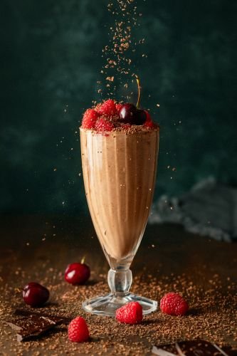 A food photographer's dream, this chocolate milkshake is artfully topped with raspberries and cherries in a tall glass. Chocolate shavings dance above, with scattered berries adding to its charm,