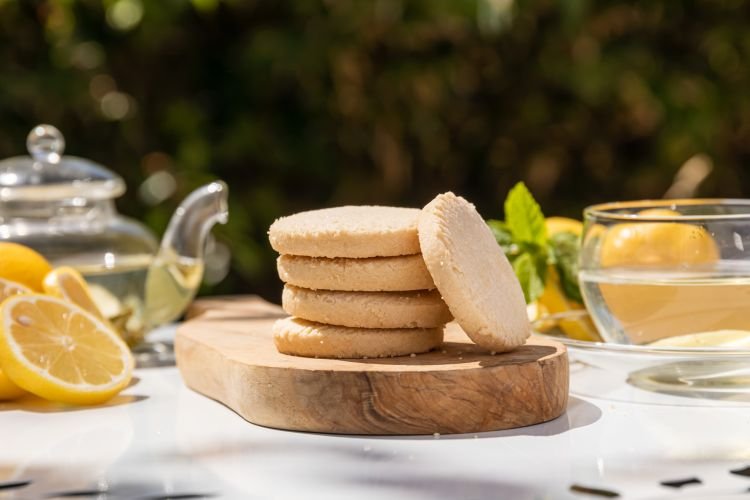 A stack of four shortbread biscuits rests on a wooden board. Surrounding the tempting treat are a glass teapot, lemon slices, a glass cup, and fresh mint leaves—an inviting scene reminiscent of serene Cornwall afternoons.