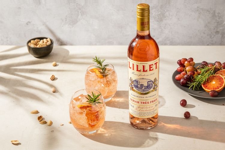 A bottle of Lillet rose vermouth stands on a bright table top next to two glasses with cocktails and a plate of grapes and grapefruit.