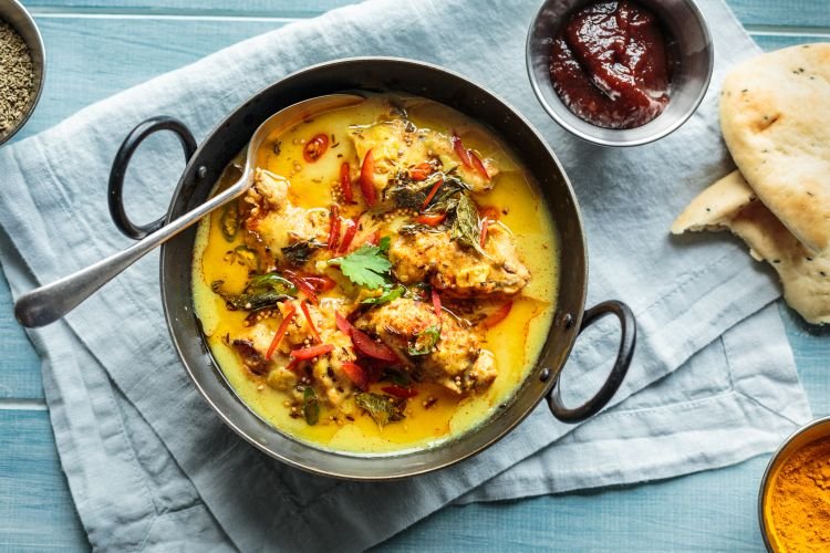 Bowl of yellow curry with chicken and herbs served with a spoon, naan bread, and a small dish of sauce on a light blue surface.