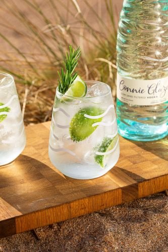 A glass with vodka, ice, lime wedges, and a sprig of rosemary sits artfully on a wooden board. Captured by a talented drinks photographer, the scene features a bottle of Connie Glaze Vodka against a background of sand and grass.