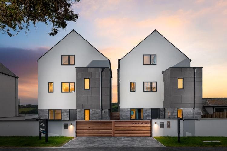 Two modern, adjacent houses with white walls and dark accents. Featuring large windows and wooden gates that provide a warm glimpse into stylish living, these homes rest elegantly under the tranquil dusk sky in Cornwall.