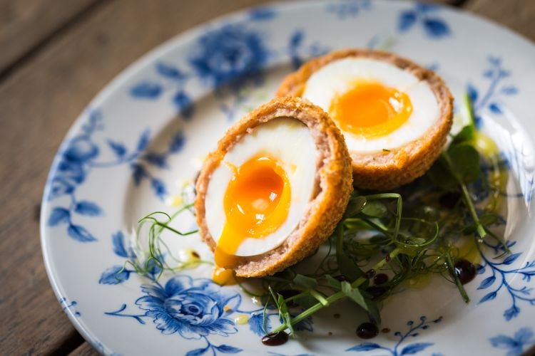 A halved soft-boiled egg wrapped in sausage and breadcrumbs is beautifully captured by a talented food photographer, presented on a blue floral plate and garnished with greens—a culinary delight inspired by the charming landscapes of Cornwall.