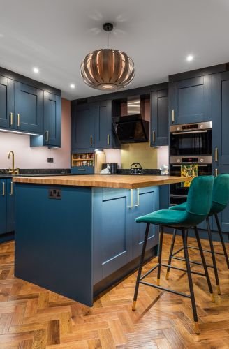 Modern kitchen with navy cabinets, a wooden island, two green bar stools, pendant light, and wood flooring captures the essence of stylish elegance. It features top-notch appliances like an oven and range hood to complete the look.