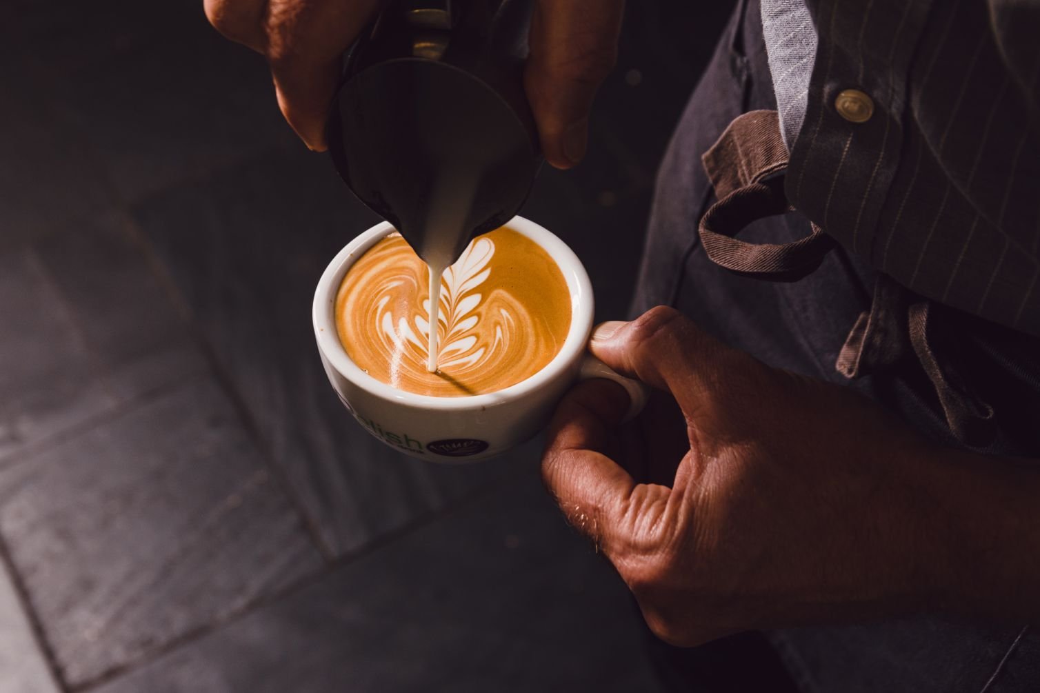 A drinks photographer in Cornwall captures the moment as a person pours steamed milk into an espresso cup, creating latte art with a stunning leaf pattern.