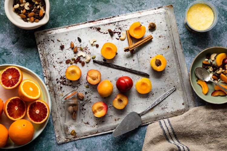 Sliced apricots, whole apricots, nuts, and cinnamon sticks on a baking tray. Nearby, vibrant bowls of oranges and mixed nuts sit alongside a blend of sliced apricots and nuts on a lush green surface.
