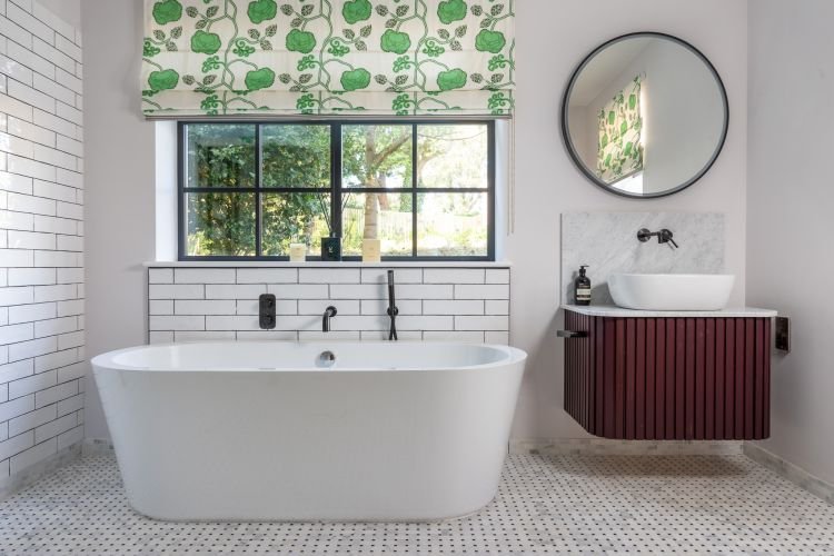 Modern bathroom captured by an interiors photographer in Cornwall, featuring a freestanding bathtub, white subway tiles, circular mirror, wall-mounted sink, and green floral window shade.