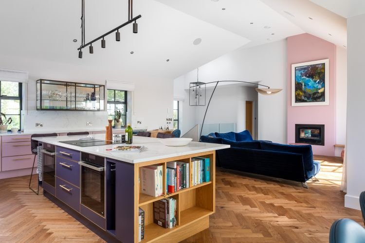 Modern open-plan kitchen and living room designed for an interiors photography spread in Cornwall, featuring a large island, bookcase, blue sofa, pink accent wall, and framed artwork.
