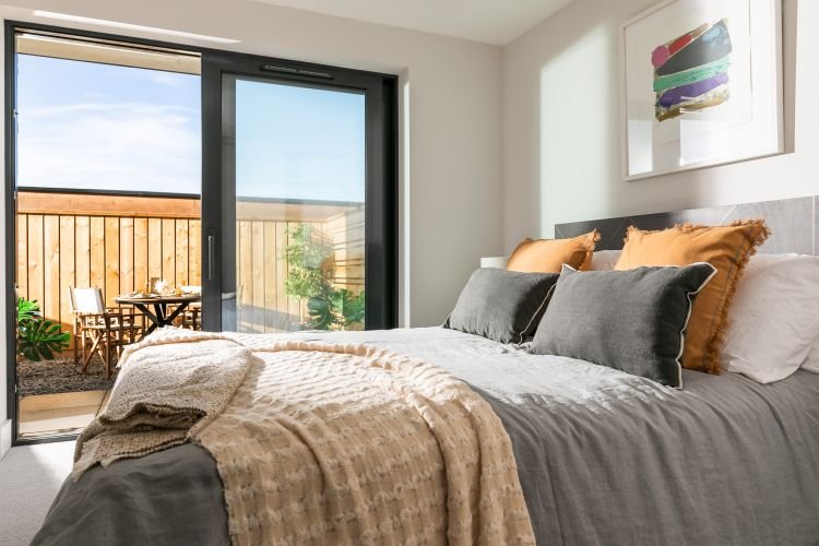 A cozy bedroom features a double bed with grey and tan bedding, two pillows, and a blanket. A sliding glass door opens to a small balcony with a table, two chairs, and a potted plant.