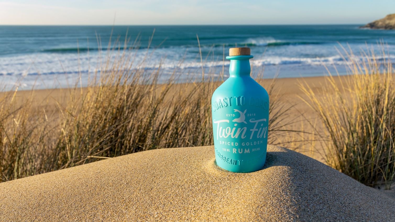 A bottle of Twinfin rum nestles in a sand dune with blue sea behind, expertly captured by a drinks photographer in Cornwall.