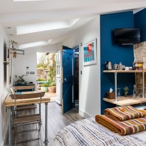 Small, modern studio with a bed, wooden dining table, open door to a patio, blue and white walls, wall-mounted TV, and stacked colorful blankets. Skylights add natural light—perfect for an interiors photographer capturing Cornwall's vibrant character.