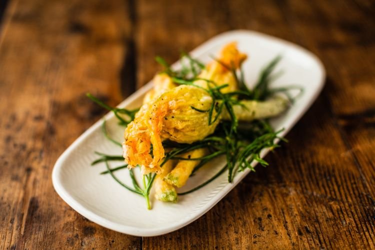 A dish of stuffed zucchini flowers garnished with herbs artfully presented on a white rectangular plate atop a rustic wooden table.