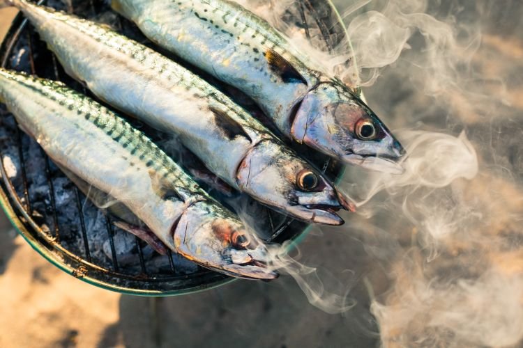 A food photographer captures the essence of beach life Cornwall with three whole mackerel sizzling on a round barbecue grill on a sandy beach.