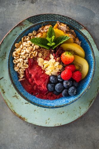 A smoothie bow is adorned with bananas, strawberries, blueberries, raspberries, sliced almonds, granola, and a mint leaf on a blue plate.