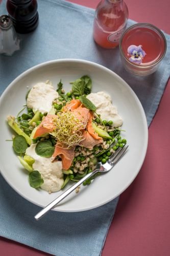 A plate of salad with salmon, greens, sprouts, and dollops of sauce sits on a white plate beside a pink drink adorned with a flower garnish on a blue cloth and pink surface.