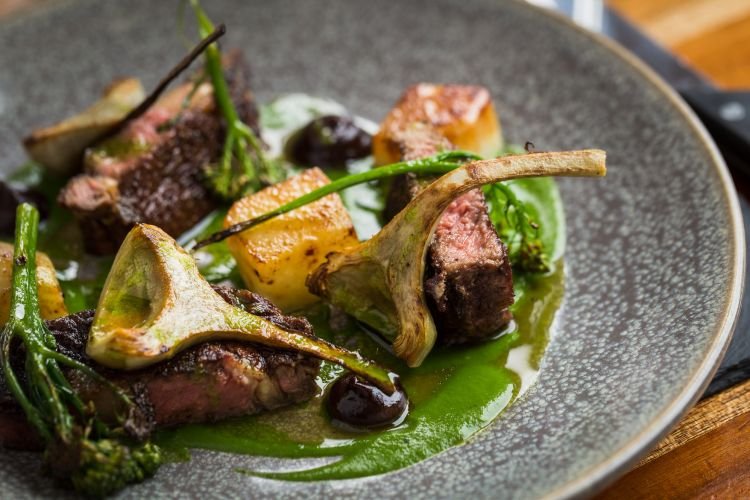 A plated dish artfully captured by a food photographer features slices of meat, artichokes, broccoli, and potatoes, garnished with a green sauce on a textured grey plate. Such visual storytelling is reminiscent of Cornwall's rich culinary landscape.