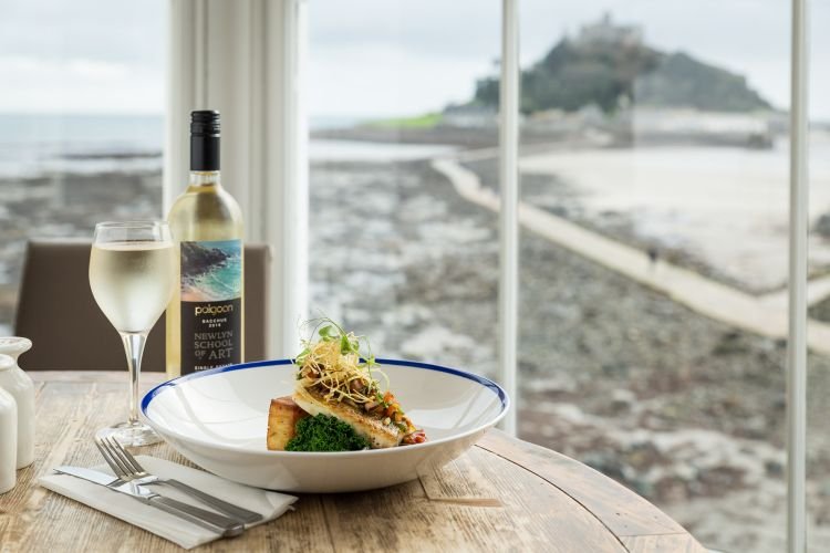 A plate of gourmet food and a glass of white wine sit elegantly on the table, perfectly capturing the seaside view through large windows overlooking St Michael's Mount in Cornwall. It's a scene worthy of any food photographer's portfolio, embodying the serene beauty of Cornwall.